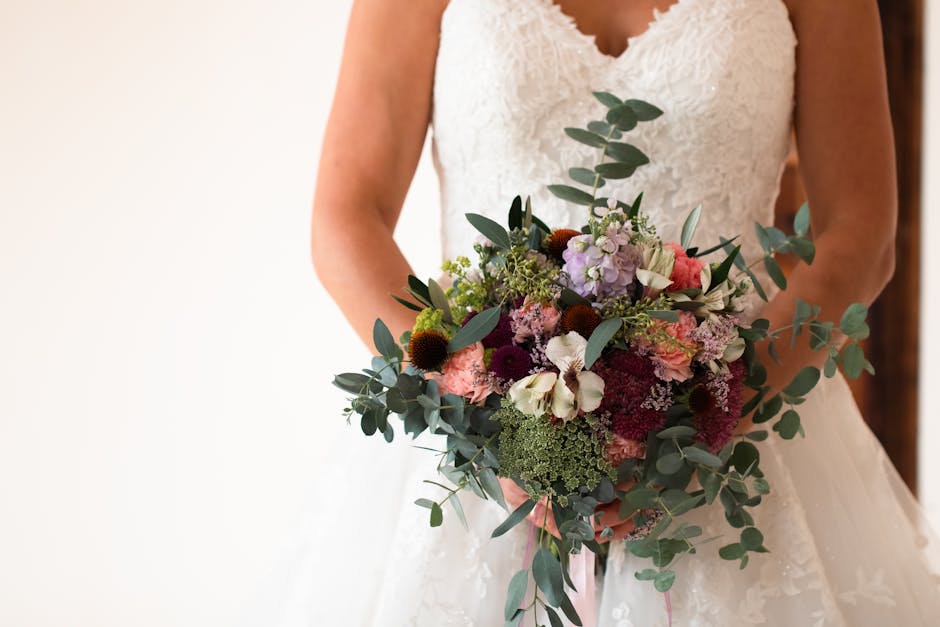 A bride in a white lace wedding dress holds a large, vibrant floral bouquet featuring a mix of fresh flowers including white orchids, pink roses, deep burgundy dahlias, purple hydrangeas, and green fo