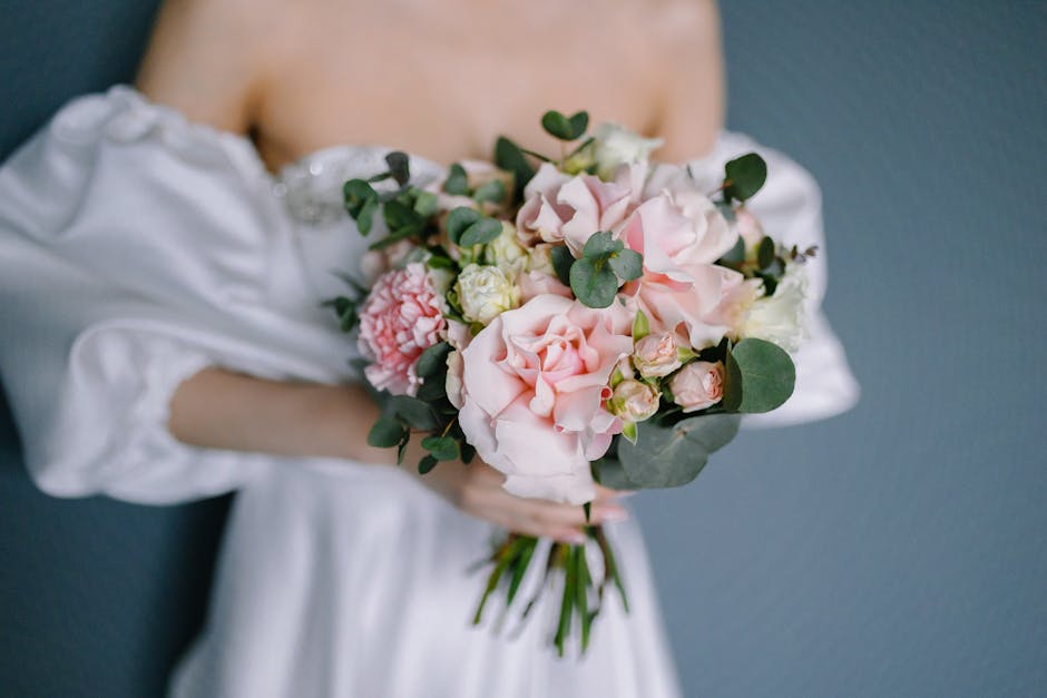 A person dressed in a white satin gown holds a fresh floral bouquet featuring soft pink roses, white lisianthus, and blush peonies, accented with eucalyptus leaves. The bouquet is arranged in a rounde