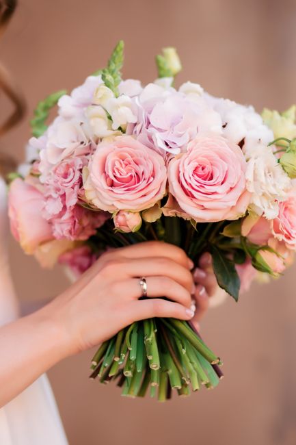 A close-up of a hand holding a bridal bouquet consisting of large, soft pink roses, white and pale pink hydrangeas, and green foliage. The stems are wrapped with a simple green wrapping, and the flowe